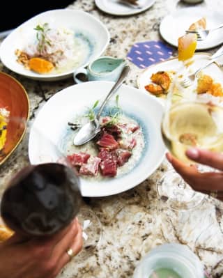 Guests raise glasses of red and white wine above a busy Tragaluz lunch, with a central plate of vibrant Nikkei tuna ceviche.