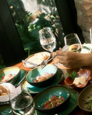 A guest reaches with a fork towards a turquoise-glazed bowl of vibrant salad, at a sun-dappled al-fresco table at Tampu.