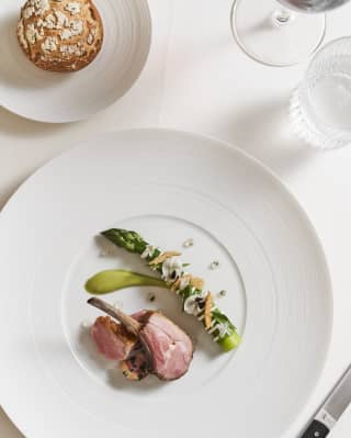 Pink lamb cutlets are artfully arranged on a large white plate with a generous asparagus spear with garnish of petals