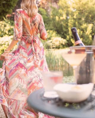 Lady in a long floaty dress strolling away from a bottle of champagne in a garden 