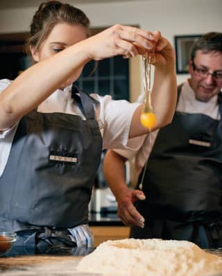 Girl in chef whites cracking an egg as a teaching chef looks on