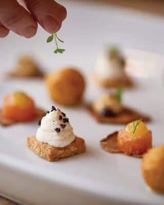 A chef carefully places a sprig of leafy garnish to a plate of colourful and enticing canapés arranged on a white plate