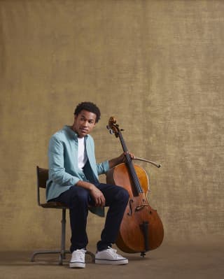 The seated young cellist, Sheku Kanneh-Mason, holds his cello and bow in his left hand as he looks off camera to the right