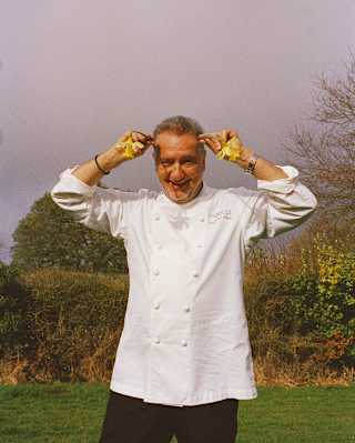 A smiling Chef Raymond Blanc poses in the garden of Manoir Aux Quatre Saisons, holding two leaves to his head like antlers.