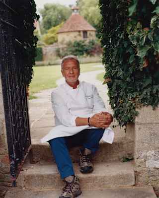 Raymond Blanc wears chef whites, blue trousers and boots as he poses for a photograph, sitting on a stone garden step.