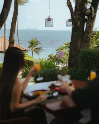 Lanterns hang from trees in Lai Thai's garden, where a couple with food and drink in soft-focus foreground gaze at the sea.