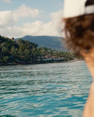 Kho Samui's palm-fringed jungle meets sparkling, aquamarine waters, seen over the shoulder of a guest on board a boat.