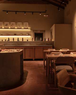 View inside Mauka's warmly-lit interior and bar, seen at the back between a focal ingredient table and row of tables.