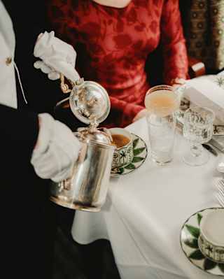 A waiter with white gloves pours tea and milk from silver lidded jugs into a William Edwards china teacup for a guest in red.