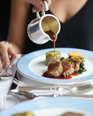 A hand pouring gravy from a silver jug onto a steak dish on a blue-rimmed plate