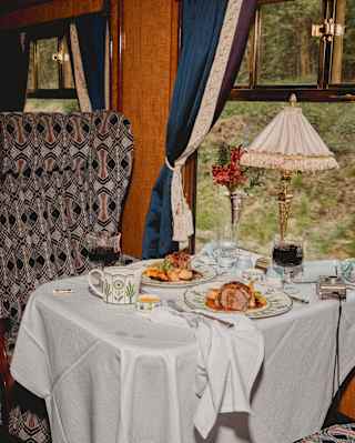 Roast dinner for two rests on a white-clothed window table with red wine and flowers, and a napkin spilling over the edge.