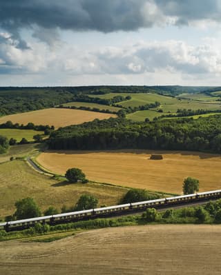 Gently rolling hills reveal a patchwork of yellow and green fields and woods. In the foreground the elegant steam train snakes