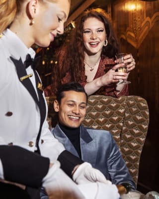 In the warm wood Zena carriage, a waitress pours champagne for a seated man as his female friend stands behind with a glass.