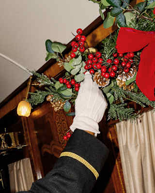 The white-gloved hand of a steward wearing a black jacket with gold braiding adjusts holly-berry carriage decoration.