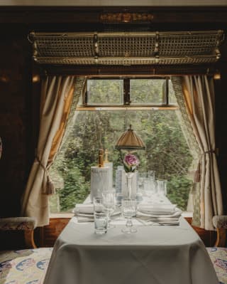 Looking at a table set for two and flanked by luxurious chairs, illuminated by the window and framed by tied-back curtains.