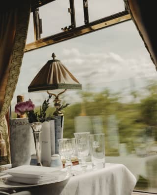 A green view rushes past the window of a dining table set for two with Champagne, bud vase and lamp, seen at eye-level.