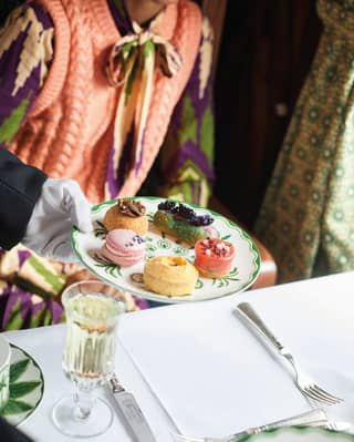 Close-up of a white-gloved hand serving a selection of afternoon tea desserts on a floral-patterned plate.