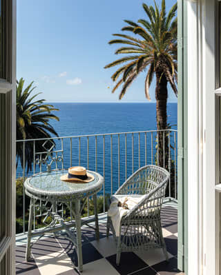 View from a bedroom to a private balcony, where a hat and glasses rest on a table and chair, with the sapphire sea beyond.