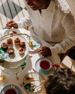Looking down on an afternoon tea with leaf-theme tableware, as two guests enjoy hot drinks and petit fours from a cake stand.