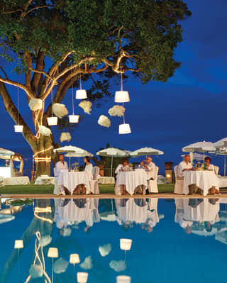 Guests in white at formal dining tables lit by hanging lanterns next to an outdoor pool