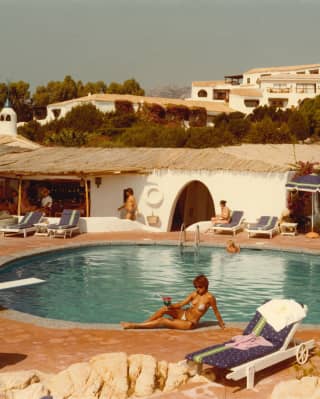 Historic honeyed Kodachrome photo of guests lounging with drinks by the sun-drenched resort pool, with a thatched bar area.