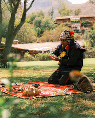 A shaman in a traditional chullo hat kneels on a red-patterned blanket laid on the lawn and covered with shells and stones.