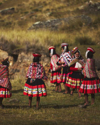 Women gather on a grassed area, dressed in vibrant traditional skirts, Manta shawls and Montera hats with sanq'apa straps.