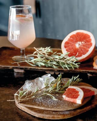 Close-up of fresh sprigs of rosemary and a sliced grapefruit beside a pink cocktail