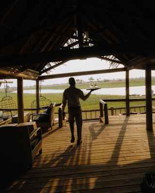 A silhouetted member of staff carries a tray with two glasses across the decked floor of the bar with a view of sunny plains.