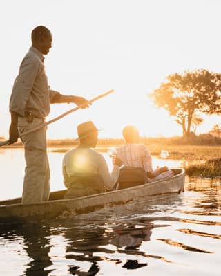 Safari guide steering two guests along a river at sunset in a traditional mokoro canoe 