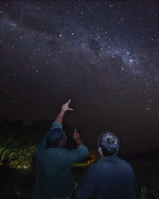 A group of star-gazers, barely visible in the dark, point at the glittery smudge of the milky way in the night sky above.