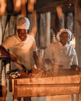 Two smiling chefs tending to food cooking on a barbecue by firelight