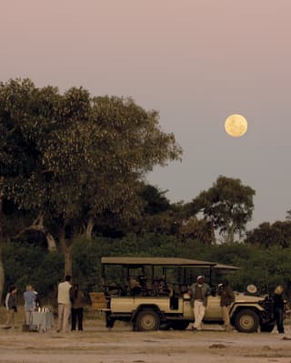 Guests surrounding a safari rover under a full moon at twilight