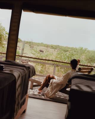 A man in a white robe and slippers reclines in a lounger on the deck of his accommodation, gazing at a passing elephant.