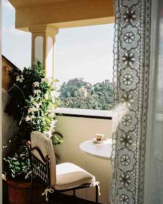 A coffee sits on a table in a warm terrace corner with a chair and blooming orange blossom, seen from behind a lace drape.