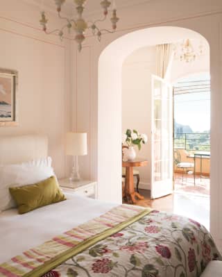 French doors lead from a furnished balcony to a sitting room and bedroom. A floral throw covers the foot of a crisp white bed