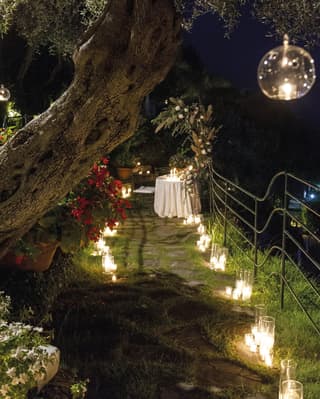 Glass globes lit with candles hang from trees. A pathway of candles lights the way through the garden to a private table