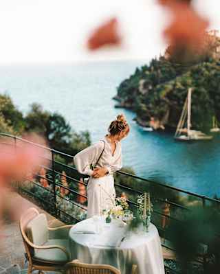 A woman in white leans on terrace railings behind a dining table for two, gazing at the dazzling bay, viewed through foliage.