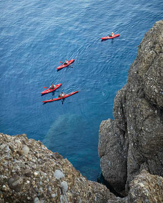 Paddlers in four kayaks explore the steep cliff coastline; their bright red kayaks small against the vastness of the sea