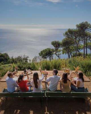 Six guests toasting with wine glasses at an outdoor table overlooking cliff top gardens