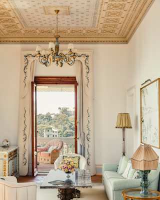 A tall, open door brings sublime views into an elegant room in pastels, with a chandelier, grey sofa and marble coffee table.