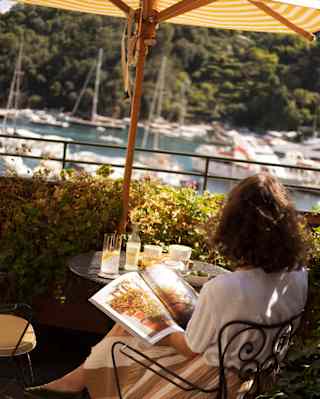 A dark-haired woman reads at a terrace table half-shaded by a parasol, overlooking the marina, seen from behind at Dav Mare.