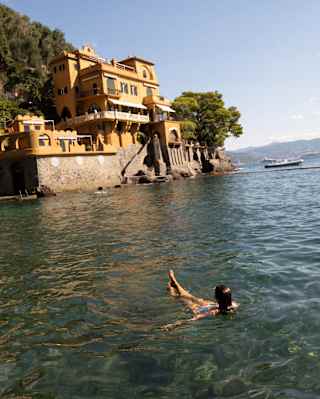 A woman floats in the clear waters of Baia Cannone facing the iconic ochre Villa Valdameri Mondadori, seen from behind.