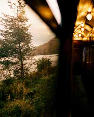 A loch with a hilly backdrop, part-obscured by a tree, is glimpsed from the window of a train corridor with glowing lights.