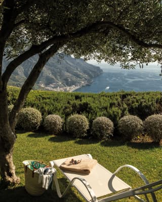 In the tranquil gardens, a basket and a sun lounger wait for a guest beneath the boughs of an olive tree, facing the coast.