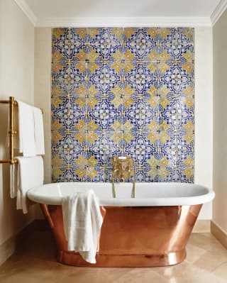 Blue and yellow geometric patterned tiles fill one wall. A free standing copper and ceramic bath reflects the marble floor