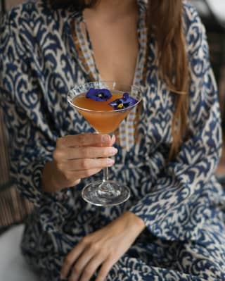 A young woman in a blue patterned kaftan holds a martini glass by the stem, her cocktail decorated with purple pansies