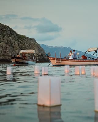 White paper box lanterns float on the calm evening sea as a young couple in a boat clink glasses on their romantic getaway