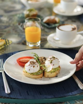 A waiter places a plate of perfect poached eggs on avocado toast on a table laden with coffee, juice and fresh fruit