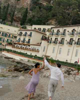 A woman in a lilac dress and male friend hold hands in the air as they walk on the beach, in an angled view from behind.